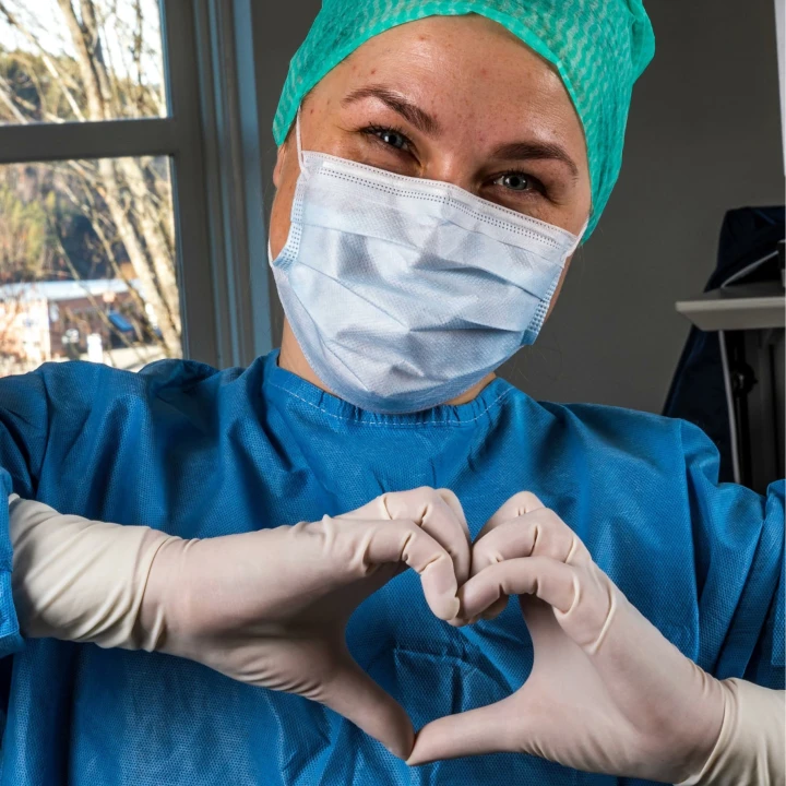 Smilende sykepleier med munnbind og sterilt tøy. Hendene er formet som et hjerte.